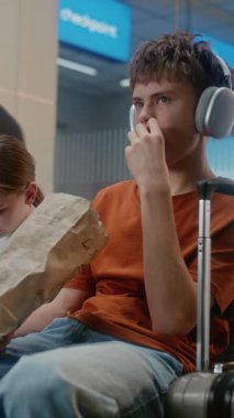 Kalabalık Uluslararası Havaalanı Terminali: Erkek ve Kız, Genç Kardeşler Abur cubur yiyor, Akıllı telefon kullanıyor, Seyahat İçin Uçuşu Bekliyor. Ayrılış Salonunda oturan çeşitli yolcular. Dikey Çekim.