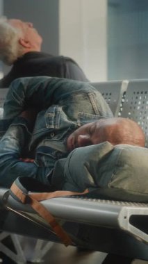 Kalabalık Uluslararası Havaalanı Terminali: Bavullu yorgun Afrikalı Amerikalı Turist ve Sırt Çantası Yatakhanedeki Koltuklarda Uyuyor. Çeşitli yolcular rötarlı uçuş bekliyor. Dikey Çekim