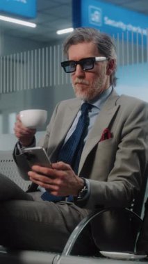 Havaalanı Terminali: Olgun İşadamı Akıllı Telefon Kullanıyor ve Kahve İçiyor, Gidiş Salonunda Bavulla Oturuyor. Girişimci uçuşu bekliyor, iş gezisine çıkıyor. Çekim, Dikey Vuruş.