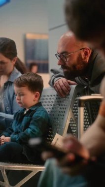 Kalabalık Havaalanı Terminali: Kulaklıklı Genç Çocuk, Akıllı Telefon Kullanıyor, Ailesi ve İki Çocuk Dijital Tablet 'te Çizgi Film İzliyor. Biniş Salonunda Uçuşu Bekleyen Çeşitli Yolcular. Dikey Çekim.