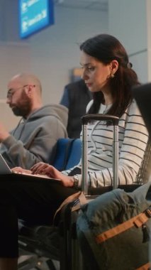 Kalabalık Uluslararası Havaalanı Terminali: Laptop 'ta Uzaktan Çalışan Kadın, E-posta Gönderen Kadın. Biniş Salonunda oturan, Digital Gadget kullanarak uçak bekleyen çeşitli yolcular. Dikey Çekim