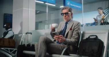 Havaalanı Terminali: Olgun İşadamı Akıllı Telefon Kullanıyor ve Kahve İçiyor, Gidiş Salonunda Bavulla Oturuyor. Girişimci uçuşu bekliyor, iş gezisine çıkıyor. Dolly shot, orta boy..