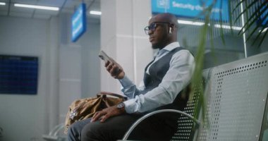 Uluslararası Havaalanı Terminali: Başarılı Girişimci Telefon Kullanıyor, E-posta Gönderiliyor. Afrika kökenli Amerikalı iş adamı, kalkış salonunda uçak bekliyor, iş gezisine çıkıyor. El bilgisayarı.