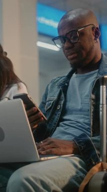 Kalabalık Uluslararası Havaalanı Terminali: Laptop 'ta Uzaktan Çalışan Afrikalı Amerikalı Adam. Biniş Salonunda oturan, Digital Gadget kullanarak uçak bekleyen çeşitli yolcular. Dikey Çekim.