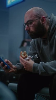 Gece Gidiş Salonu: Burger Yiyen Adam ve Kaydıran Telefon Kalabalık Uluslararası Havaalanı Terminalinde Oturuyor. Yorgun İnsanlar ve Yolcular Gecikmiş Uçak Uçuşunu Bekliyor. Dikey Çekim.