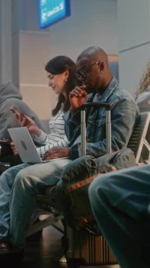 Kalabalık Uluslararası Havaalanı Terminali: Çeşitli İnsanlar, Ayrılış Salonunda oturan Yolcular, Uçak Uçuşunu Bekliyor, Telefonları Kullanıyor. İnternette çalışan Afrikalı Amerikalı adam. Dikey Çekim.