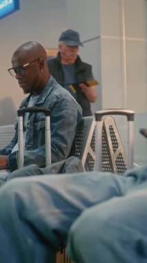 Kalabalık Uluslararası Havaalanı Terminali: Ayrılış Salonunda Çeşitli İnsanlar ve Yolcular. Bilgisayarda çalışan Afro-Amerikalı Adam Otururken ve Uçak Uçuşunu beklerken. Dikey Çekim.