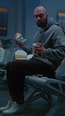 Kalabalık Uluslararası Havaalanı Terminali: Burger Yiyen ve Gece Oturan Kahve İçen Adam. Yorgun İnsanlar ve Yolcular Gecikmiş Uçak Uçuşunu Bekliyor. Dikey Çekim.