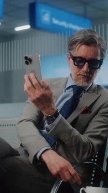 Havaalanı Terminali: Akıllı Telefonla Konuşan Başarılı Girişimci Video Sohbeti, İçen Kahve. İş adamı kalkış salonunda uçuşu bekliyor, iş gezisine çıkıyor. Ağır çekim. Dikey Çekim.