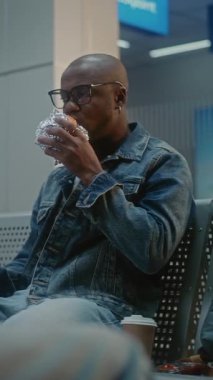 Kalabalık Uluslararası Havaalanı Terminali: Afro-Amerikan Adam Fast Food yiyor ve akıllı telefon kullanıyor. Çeşitli Yolcular Gecikmiş Uçak Uçuşunu Bekliyor. Dikey Çekim.