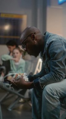 Kalabalık Uluslararası Havaalanı Terminali: Afro-Amerikan Adam Burger Yiyor ve Kaydırma Telefonu Gecikmeli Uçuşu Bekliyor. Ayrılış Salonunda oturan çeşitli insanlar ve yolcular. Dikey Çekim.