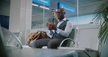 Uluslararası Havalimanı Terminali: Akıllı Telefon Kullanan Afrikalı Amerikalı İşadamı, E-İş Yapıyor. Erkek Girişimci Ayrılış Salonunda Uçak Uçuşunu Bekliyor, İş Gezisine Gidiyor. El bilgisayarı.