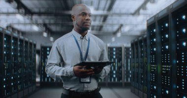 Sunucu Raflı Büyük Veri Merkezi: Profesyonel Afro-Amerikan Uzmanı Tablet Bilgisayar Kullanıyor, Sunucuları Etkinleştiriyor, Depo 'da Duruyor. Bilgi Teknolojisi ve Siber Güvenlik. Orta Çekim.