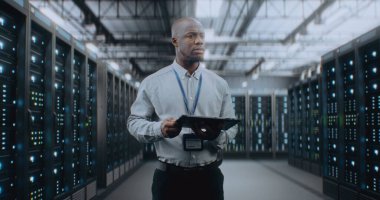 Sunucu Raflı Büyük Veri Merkezi: Profesyonel Afro-Amerikan Uzmanı Tablet Bilgisayar Kullanıyor, Sunucuları Etkinleştiriyor, Depo 'da Duruyor. Bilgi Teknolojisi ve Siber Güvenlik. Orta Çekim.
