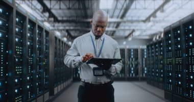 Sunucu Raflı Büyük Veri Merkezi: Profesyonel Afro-Amerikan Uzmanı Tablet Bilgisayar Kullanıyor, Sunucuları Etkinleştiriyor, Depo 'da Duruyor. Bilgi Teknolojisi ve Siber Güvenlik. Orta Çekim.