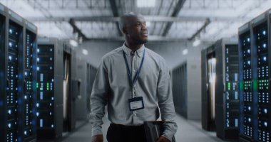 Sunucu Raflı Büyük Veri Merkezi: Tablet Bilgisayar Deposunda Yürüyen Profesyonel Afro-Amerikan Uzmanı. İnternet Sunucuları, Bilgi Teknolojisi, Siber Güvenlik ve Haberleşme.
