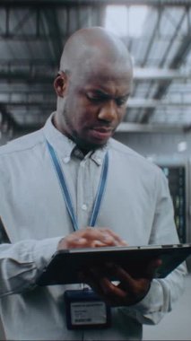 Sunucu Raflı Büyük Veri Merkezi: Profesyonel Afro-Amerikan Uzmanı Tablet Bilgisayar Kullanıyor, Sunucuları Etkinleştiriyor, Depo 'da Duruyor. Bilgi Teknolojisi ve Siber Güvenlik. Dikey Çekim.