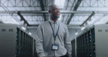 Sunucu Raflı Büyük Veri Merkezi: Tablet Bilgisayar Deposunda yürüyen, Kameraya bakan profesyonel Afro-Amerikan Uzmanı. İnternet Sunucuları, Bilgi Teknolojisi ve Siber Güvenlik.