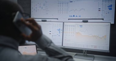 Cep Telefonu Konuşan Finansal Analistin Kapanışı, Bilgisayarda Çoklu İzleyici Çalışma İstasyonu ile Gerçek Zamanlı Borsa Çizelgeleri İzleniyor. Afrikalı Amerikalı İşadamı Ofiste Çalışıyor.