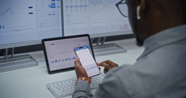 Çevrimiçi Ticaret Uygulaması Kullanılarak Finansal Analizci Gerçek Zamanlı Borsa Çizelgelerini, Akıllı Telefon ve Dijital Ekranları Kontrol Ediyor. Afrikalı Amerikalı İşadamı Yatırım Bankası Ofisi 'nde çalışıyor. Kapat..