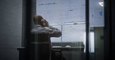 Finansal Analizci Telefonda Konuşuyor, Bilgisayar üzerinde Multi-Monitor Çalışma İstasyonu ile çalışıyor, Gerçek zamanlı borsa ve borsa grafiklerini analiz ediyor. Afrikalı Amerikalı İşadamı Geceleri Ofiste Çalışıyor.