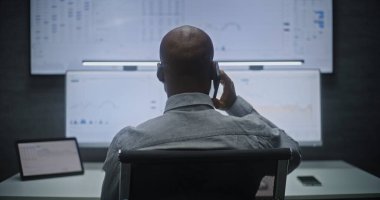 Multi-Monitor Workstation ile bilgisayar üzerinde çalışan Finansal Analizci, Gerçek zamanlı Borsa Çizelgelerini İzleyen, Telefonda Konuşan. Afrikalı Amerikalı İşadamı Geceleri Ofiste Çalışıyor.