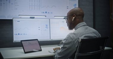Finansal Analizci Gerçek Zamanlı Hisse Senetlerini İzliyor, Bilgisayarda Çoklu Monitör Çalışma İstasyonu ile Piyasa Tablolarını Değiştiriyor, Kablosuz Kulaklıkla Konuşuyor. Afrikalı Amerikalı İşadamı Geceleri Ofiste Çalışıyor.