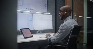 Finansal Analizci Kablosuz Kulaklıkla Konuşuyor, Gerçek zamanlı Borsa İzleme, Çoklu Monitör Çalışma İstasyonu ile Bilgisayarda Piyasa Tabloları Değiştirme. Afrikalı Amerikalı İşadamı Geceleri Ofiste Çalışıyor.