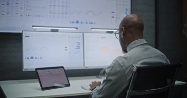 Finansal Analizci Gerçek Zamanlı Hisse Senetlerini İzliyor, Bilgisayarda Çoklu Monitör Çalışma İstasyonu ile Piyasa Tablolarını Değiştiriyor, Kablosuz Kulaklıkla Konuşuyor. Afrikalı Amerikalı İşadamı Geceleri Ofiste Çalışıyor.