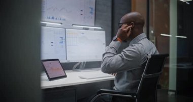 Finansal Analizci Kablosuz Kulaklıkla Konuşuyor, Gerçek zamanlı Borsa İzleme, Çoklu Monitör Çalışma İstasyonu ile Bilgisayarda Piyasa Tabloları Değiştirme. Afrikalı Amerikalı İşadamı Geceleri Ofiste Çalışıyor.