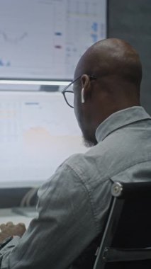 Finansal Analizci Gerçek Zamanlı Hisse Senetlerini İzliyor, Bilgisayarda Çoklu Monitör Çalışma İstasyonu ile Piyasa Tablolarını Değiştiriyor, Kablosuz Kulaklıkla Konuşuyor. Ofisteki Afrikalı Amerikalı İş Adamı. Dikey Çekim.
