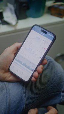 Gerçek zamanlı borsa çizelgelerini kontrol eden adam akıllı telefon kullanıyor. Çalışanların atölyede çalışma saatleri sırasında yatırım yapması, İş ve Çevrimiçi Ticaret 'i birleştirmesi. Yakın çekim. Dikey Çekim.