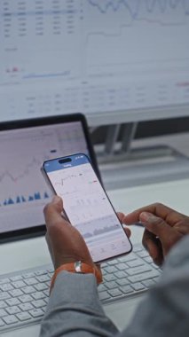 Finansal Analizci, Smartphone 'da Gerçek Zamanlı Borsa Çizelgelerini ve Ticari Uygulamayı kullanarak Dijital Ekranları Kontrol Ediyor. Afrikalı Amerikalı İşadamı Bankada çalışıyor. Yakın çekim. Dikey Çekim.