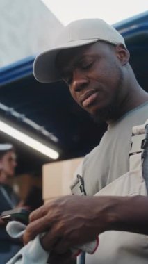 Lojistik Deposunun dışında Afro-Amerikan Yükleyici Akıllı Telefon Kullanıyor, Şaşırmış Bakışlı Kameraya Bakıyor. Çeşitli işçiler teslimat için karton kutularla kargo kamyonu yüklüyor. Dikey Çekim