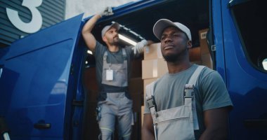 Lojistik Dağıtım Deposu 'nun önünde dikilen, gülümseyen ve kameraya bakan iki farklı üniformalı işçi ekibi. E-ticaret çevrimiçi siparişlerle dolu karton kutularla dolu bir nakliye kamyonu..