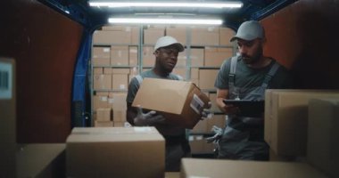 Lojistik Perakende Deposu 'nun dışında muhtelif çalışanlar karton kutularla kargo kamyonu boşaltıyorlar. Dijital Tablet kullanarak Yönetici Tarayıcı Paketleri. Çevrimiçi Siparişler, Satın Almalar ve E-Ticari Mallar.