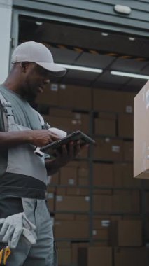 Çeşitli işçiler E-Ticaret Çevrimiçi Siparişleri ile karton kutuları Lojistik Deposuna götürüyor. Tablet Bilgisayar kullanarak, Barkod Tarayıcı ile Afrikalı Amerikalı Yönetici Paketleri tarıyor. Dikey Çekim.