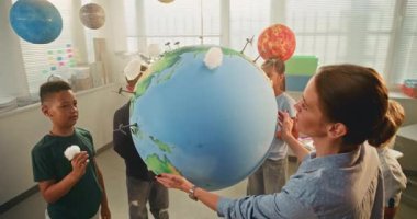 Bilim Dersi: Çok Irklı Sınıfta Akıllı Çocuklara Yenilenebilir Enerji Eğitimi Yapan Kadın Öğretmen. İlkokul çocukları Dünya Modeline El Yapımı Bulutlar Ekliyor. BTMM Eğitim. Yüksek Açı