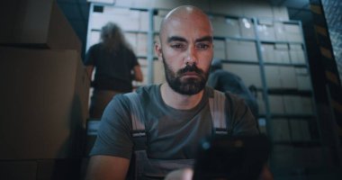 Yorgun Yükleyici Oturma ve Akıllı Telefon Kullanma, Gece Lojistik Deposunda Sıkı Çalışma Gününden sonra Dinlenme. Arka planda, çeşitli işçiler E-ticaret ürünleriyle karton kutular taşıyorlar. El bilgisayarı.