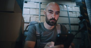 Yorgun Yükleyici Oturma ve Akıllı Telefon Kullanma, Gece Lojistik Deposunda Sıkı Çalışma Gününden sonra Dinlenme. Arka planda, çeşitli işçiler E-ticaret ürünleriyle karton kutular taşıyorlar. El bilgisayarı.