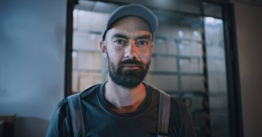 Gece Lojistik Perakende Deposu 'nun dışında Üniformalı Erkek İşçi Portresi Kameraya Bakıyor, Teslimat Servisi' nde çalışıyor. Yükleyiciler arka planda karton kutular taşıyor. Görüntüyü Kapat.