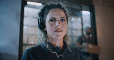 Gece Lojistik Dağıtım Deposu 'nun dışında: Kameraya Bakan Kadın Kulaklık Giyen, Teslimat Servisi' nde çalışan. Yükleyiciler arka planda paket taşıyorlar. Kapat..