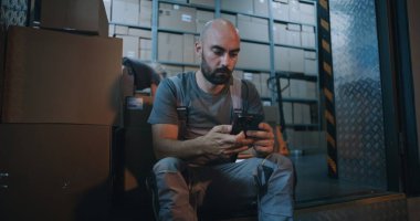Çevrimiçi sipariş ile karton kutu taşıyan yorgun işçi, mola veren, akıllı telefon kullanan. Arka planda, geceleri Lojistik Dağıtım Deposunda çalışan çeşitli çalışanlar var. Kargo Servisi.