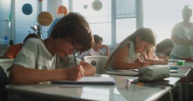 İlkokul öğrencileri Sınav Yazıyor ya da Not Defterlerinde Görev Yapıyor. Modern Sınıfta Sınav Sürecinde Masalar Arasında Yürüyen Bayan Öğretmen, Genç Kızları ve Oğlanları Kontrol Etmek. Doli Vuruşu.