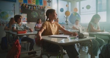 İlkokul çocukları masada oturuyor, öğretmenden ders dinliyor, doğru cevap vermek için ellerini kaldırıyor. Modern Sınıfta Coğrafya veya Bilim Öğrenen Genç Erkekler ve Kızlar Grubu.