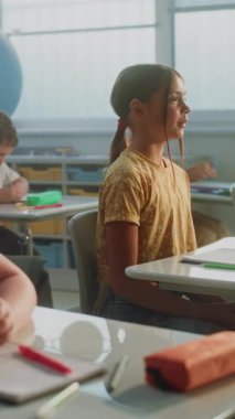 İlkokul çocukları masada oturuyor, öğretmenden ders dinliyor, doğru cevap vermek için ellerini kaldırıyor. Sınıfta Coğrafya Öğrenen Genç Erkekler ve Kızlar Grubu. Dikey Çekim.