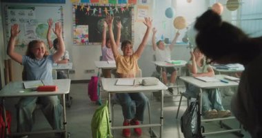 Bayan öğretmen ilkokul çocuklarına komik etkileşimli ders veriyor. Masada oturan, el kaldıran, dinamik sınıf etkinliği sırasında öğretmenle etkileşim kuran mutlu genç kızlar ve oğlanlar..