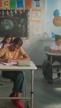 İlkokul öğrencileri Sınıfta Yenilenebilir Enerji Kaynakları Sınavı Yazıyor. Masaların Arasında Yürüyen Bayan Öğretmen, Okul Sınavı Sürecinde Genç Kız ve Oğlanları Kontrol Etmek. Dikey Çekim.