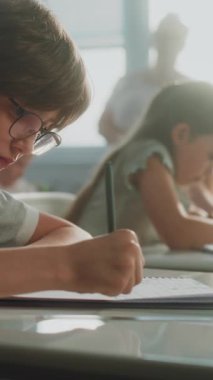 İlkokul çocukları, yazma okulu sınavı, not defterlerinde görevler. Masalar Arasında Yürüyen Kadın Öğretmen, Modern Sınıfta Sınav Sürecinde Çeşitli Çocuklar Kontrol Grubu. Dikey Çekim.