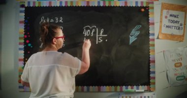 Tebeşir tahtasında yazan, Matematik dersi sırasında ilkokul çocuklarına çarpma ve bölmeyi açıklayan kadın öğretmenin geçmişe bakışı. Modern Sınıfta Matematik Sınıfı. Sabit Çekim.